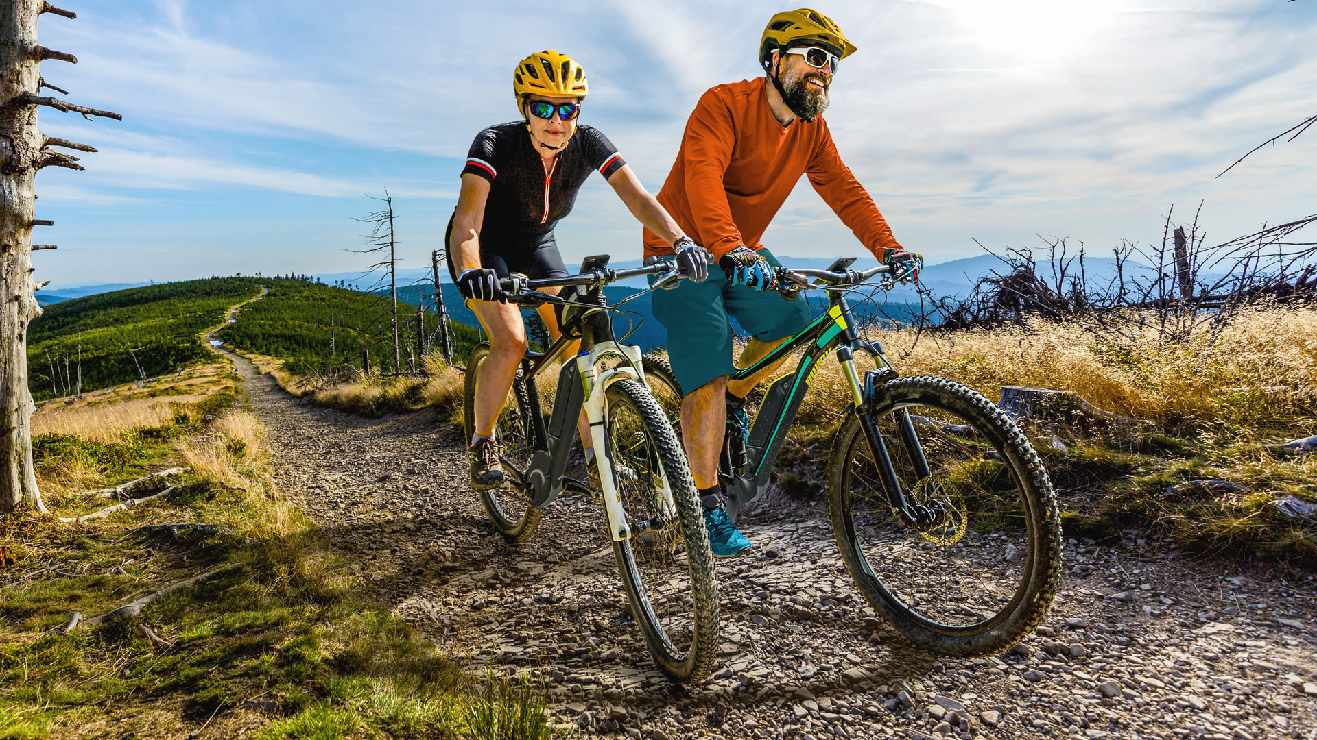 Zwei Mountainbiker fahren auf einem schmalen Pfad durch eine hügelige Landschaft.