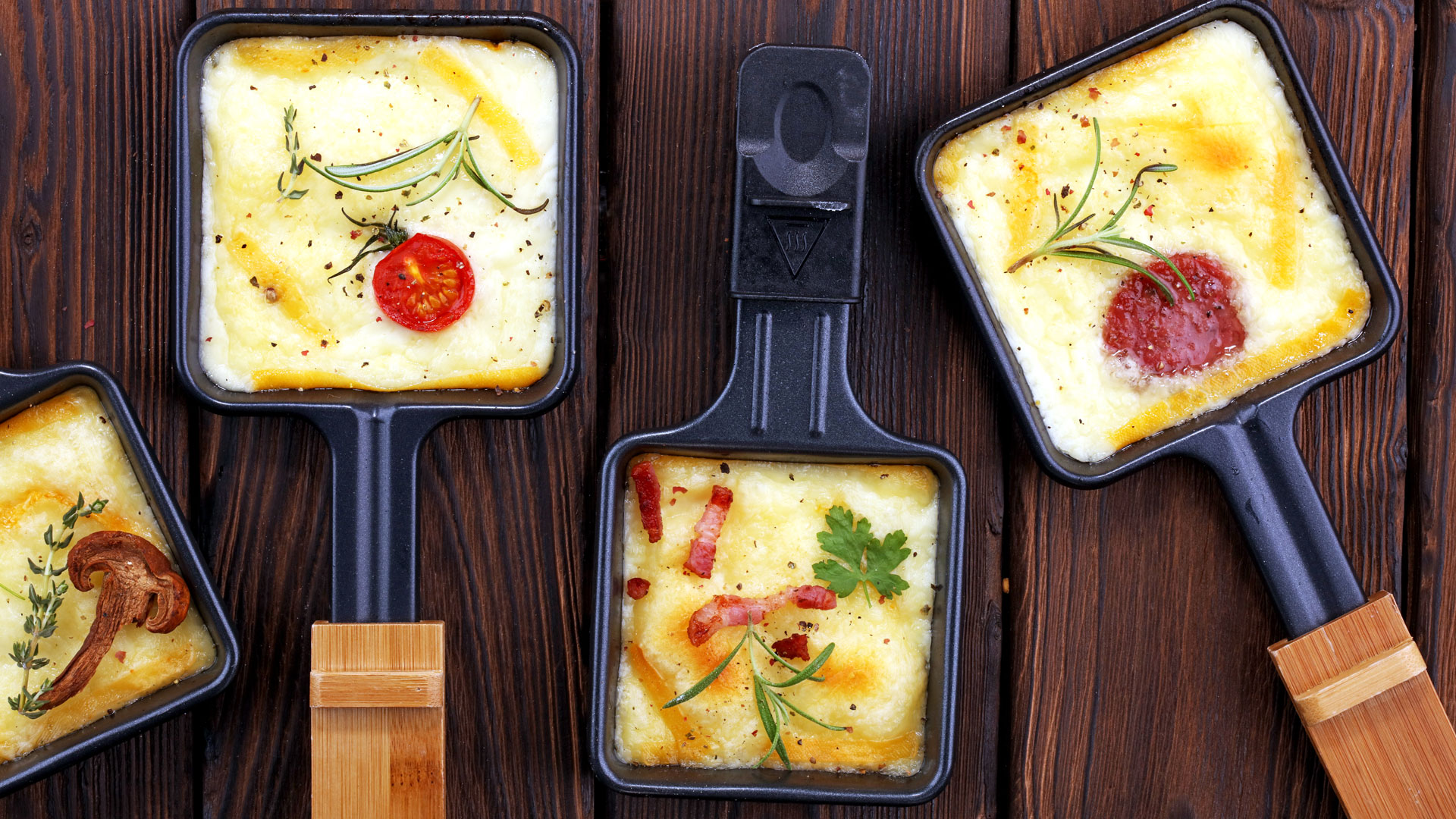 Vier kleine Pfännchen mit überbackenen Eierspeisen und verschiedenen Zutaten wie Tomaten, Speck und Pilzen.
