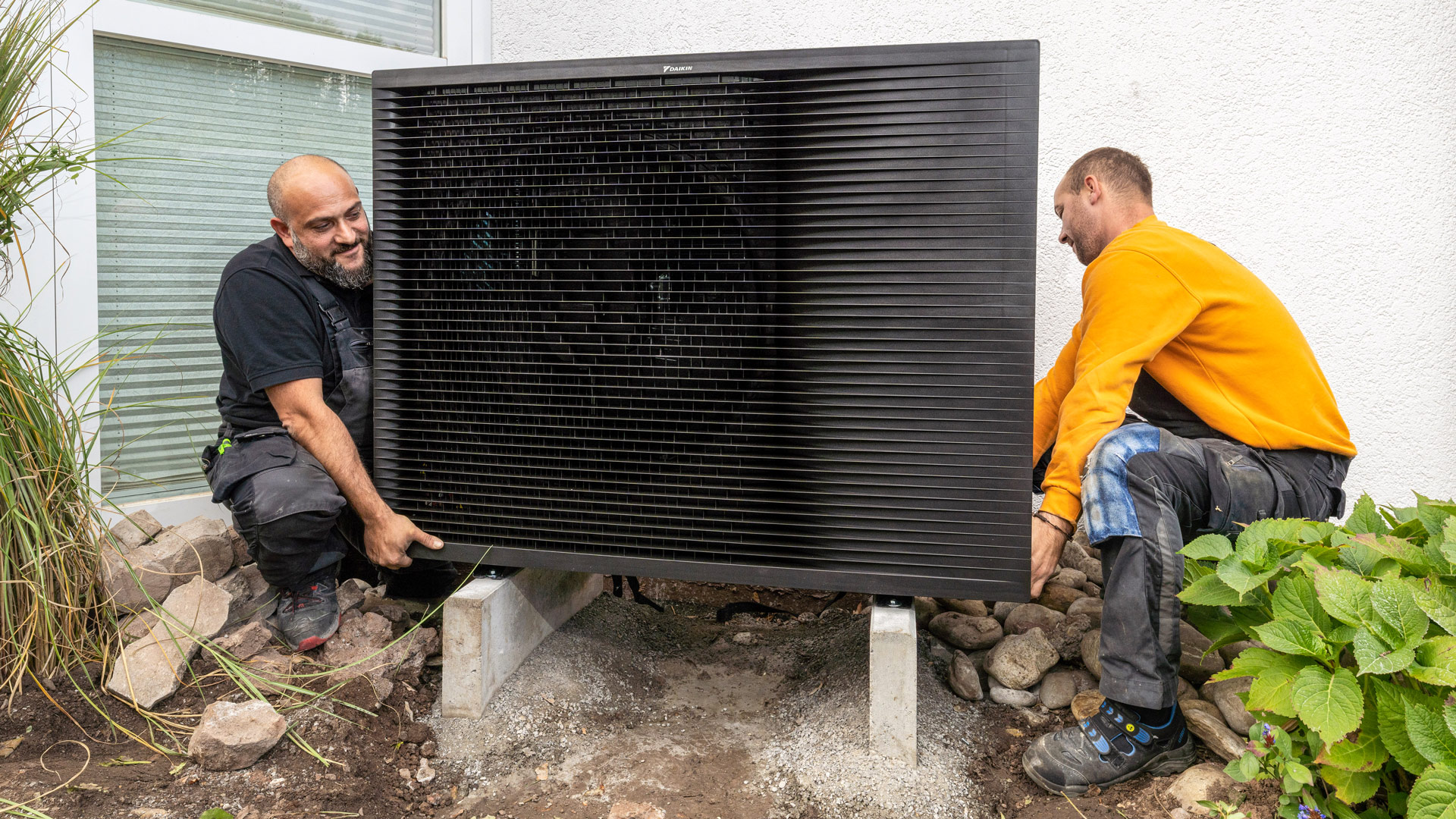 Zwei Handwerker installieren eine Wärmepumpe im Garten.