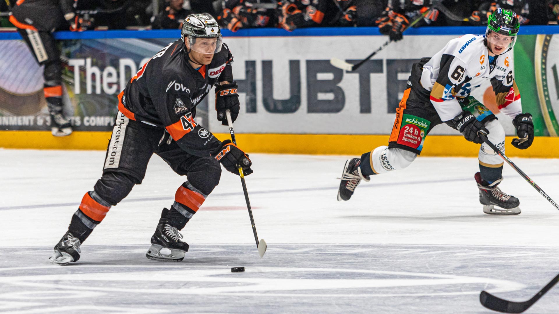 Eishockeyspieler mit schwarzem Trikot und Helm im Spiel, verfolgt von Gegenspieler mit blauem Trikot.