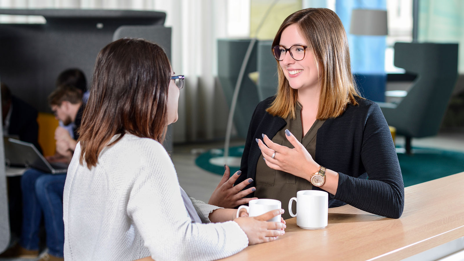 Zwei Frauen sitzen an einem Tisch und unterhalten sich. Die Frau links hält eine Tasse, die Frau rechts gestikuliert.