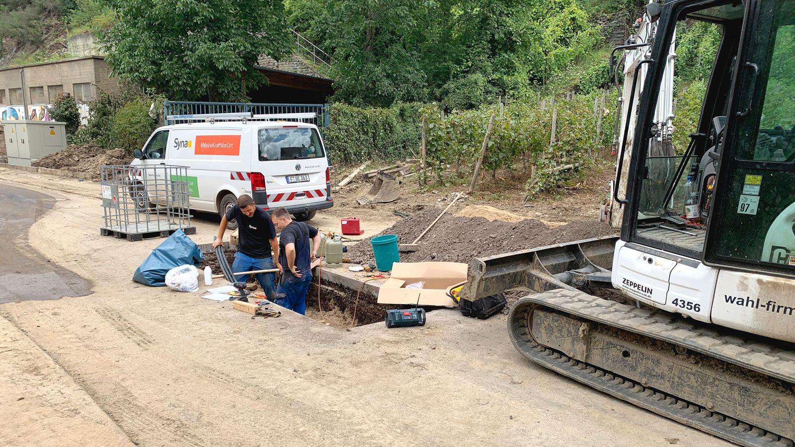 Zwei Personen arbeiten an einer Baustelle, neben einem Bagger und einem weißen Transporter mit Aufschrift "Syna".