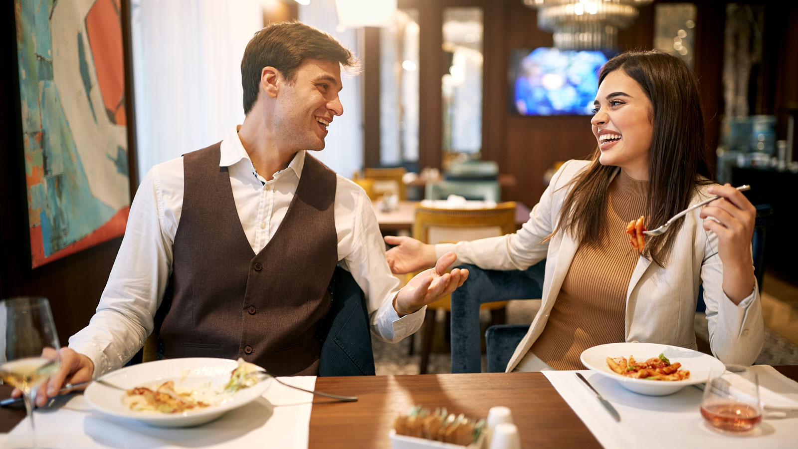 Zwei Personen sitzen an einem Tisch und essen Pasta. Der Mann gestikuliert, die Frau lächelt.