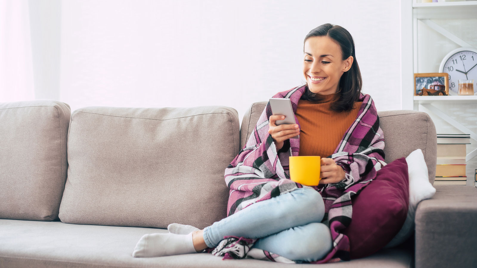 Frau sitzt auf Sofa, liest Smartphone und hält Tasse.