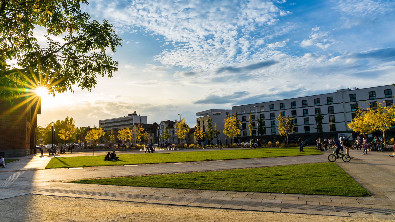 Grünfläche mit Personen und Gebäuden im Hintergrund, sonniger Tag.