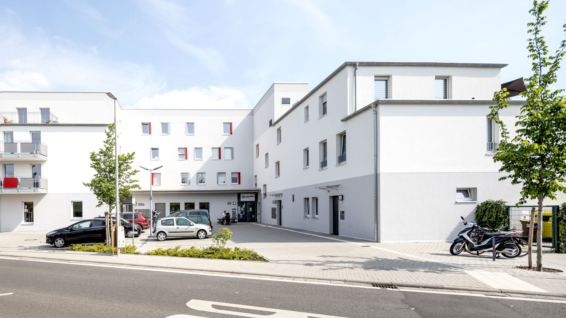 Mehrstöckiges Wohnhaus mit Parkplätzen und Motorrollern davor.