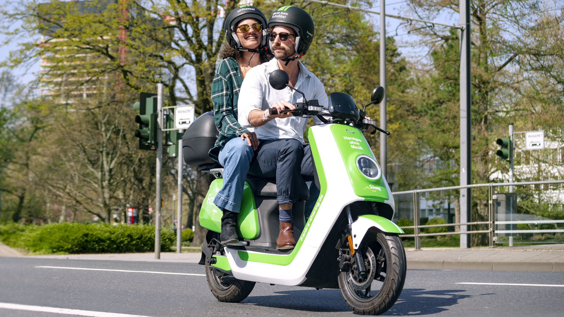Zwei Personen auf grünem Elektroroller, beide tragen Helme.