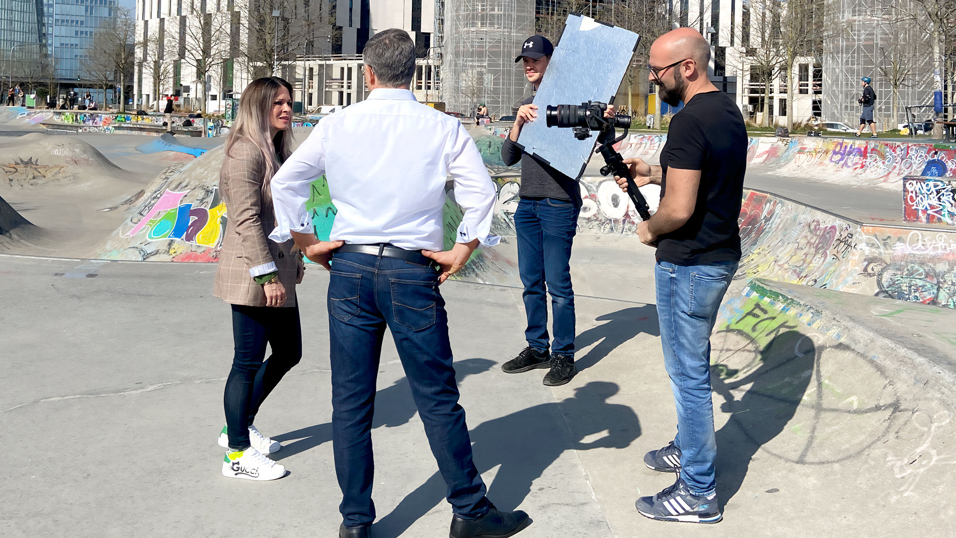 Drei Personen filmen vor einer Skateanlage. Links eine Frau in beigem Blazer, in der Mitte ein Mann im Hemd und Jeans, rechts ein Mann mit Kamera.
