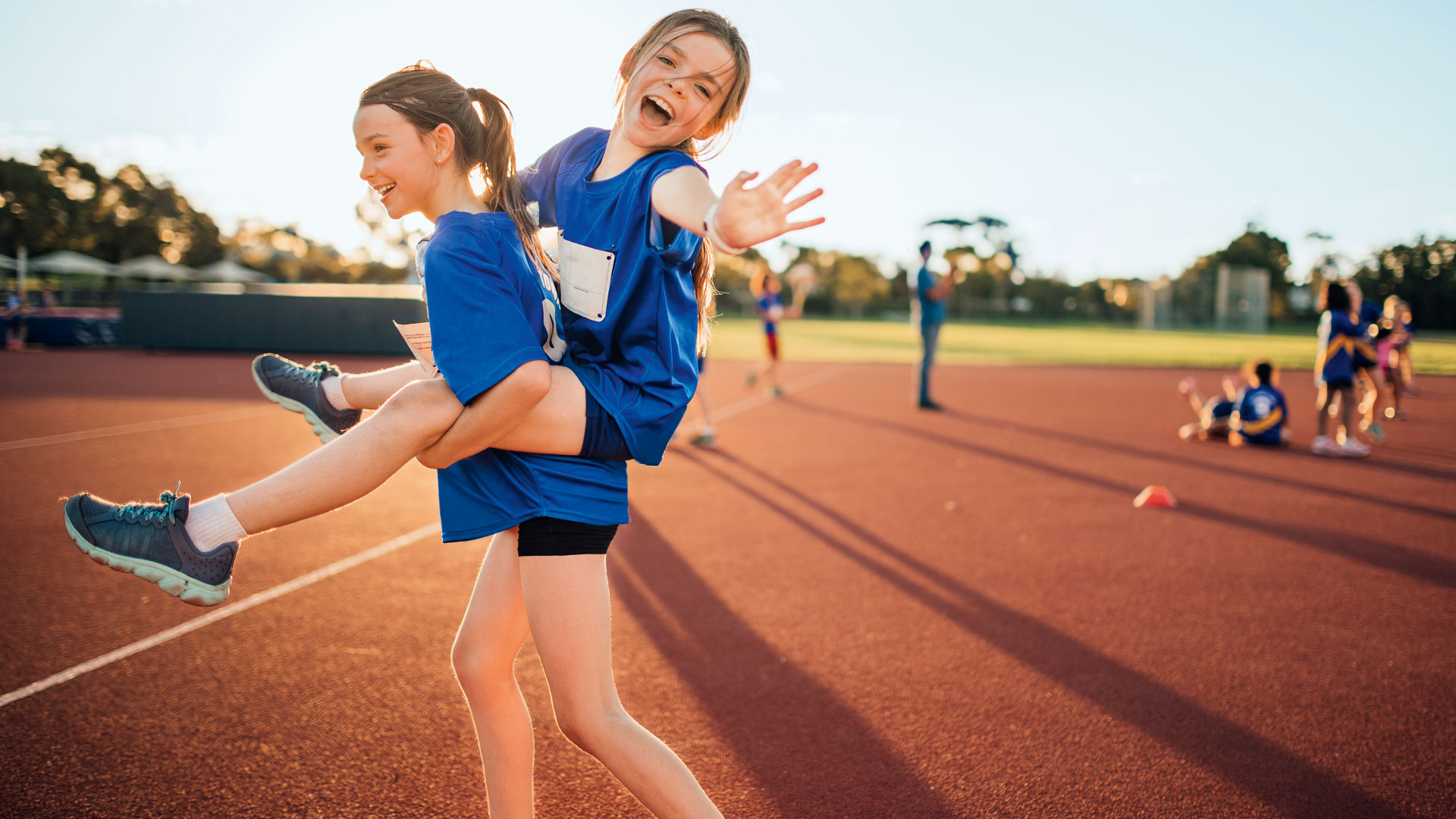 Zwei Mädchen laufen auf einer roten Tartanbahn. Das eine Mädchen trägt das andere auf den Schultern.