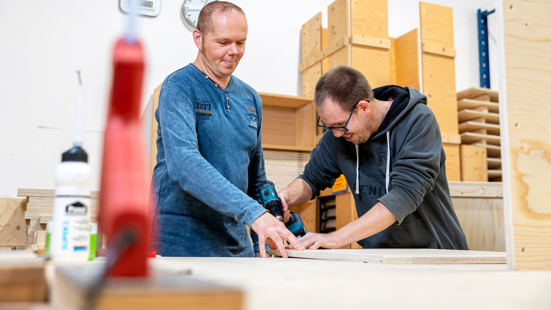 Zwei Handwerker bearbeiten ein Holzbrett mit Elektrowerkzeug.