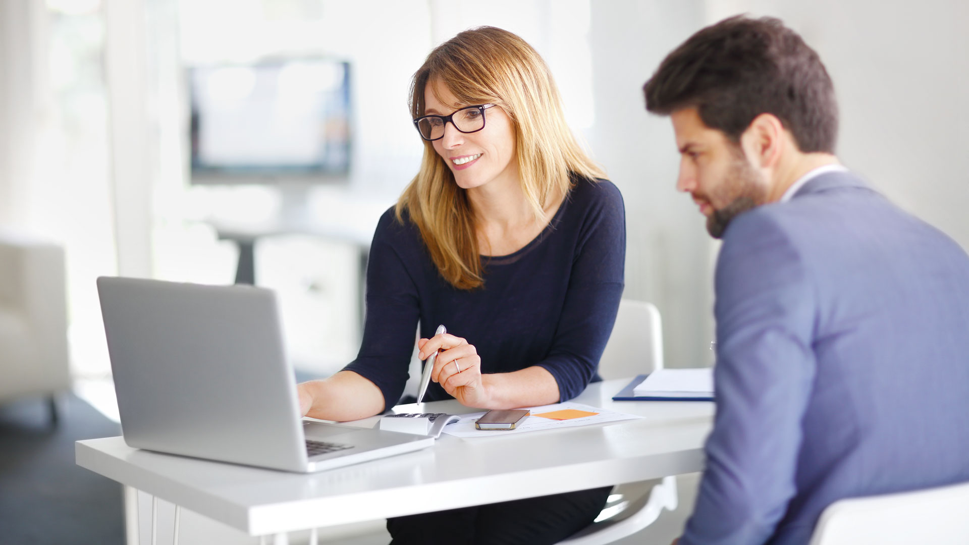 Zwei Personen sitzen an einem Tisch, eine Frau tippt auf einem Laptop, der Mann schaut zu.