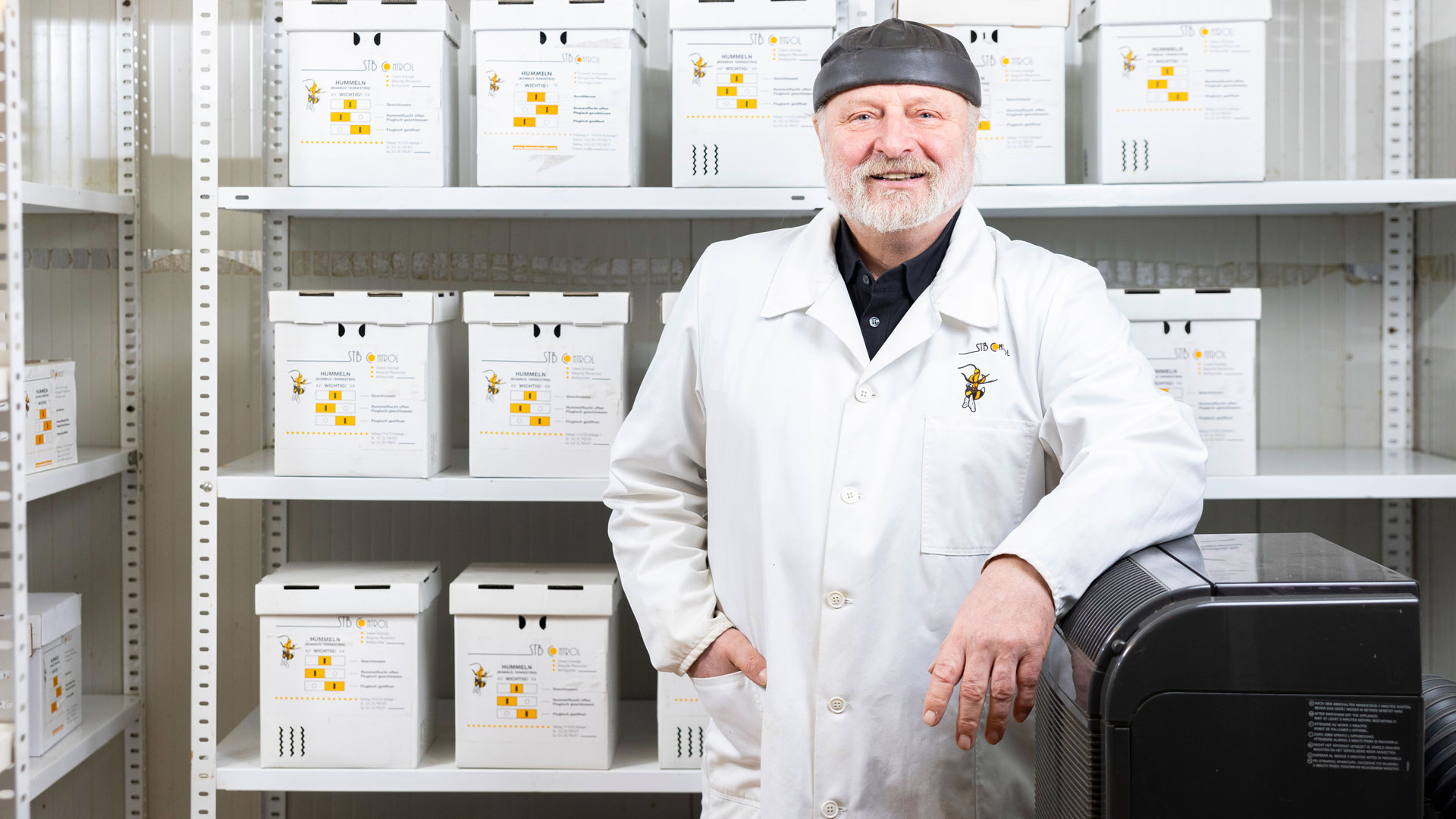 Mann in weißem Kittel vor Regalen mit beschrifteten Ordnern.