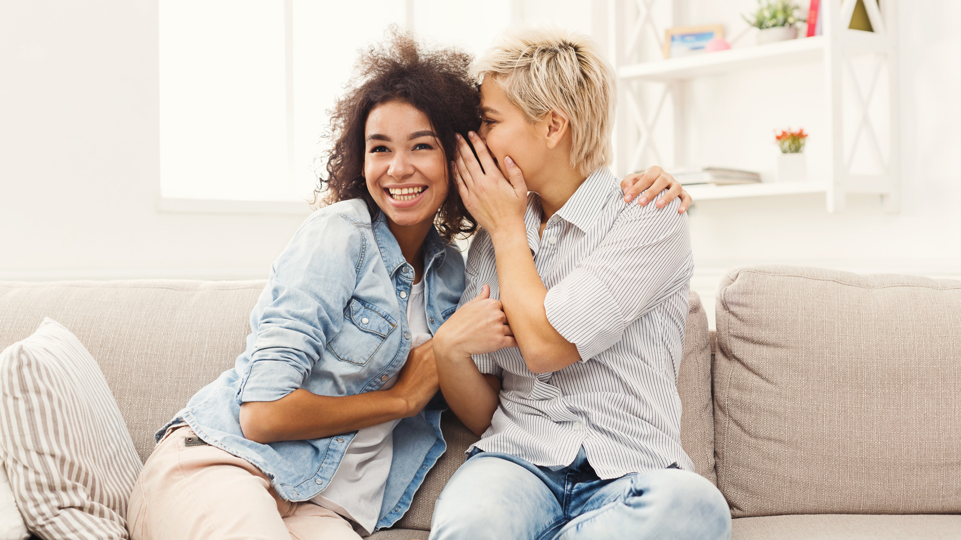 Zwei Frauen sitzen auf einem Sofa und flüstern sich etwas ins Ohr.