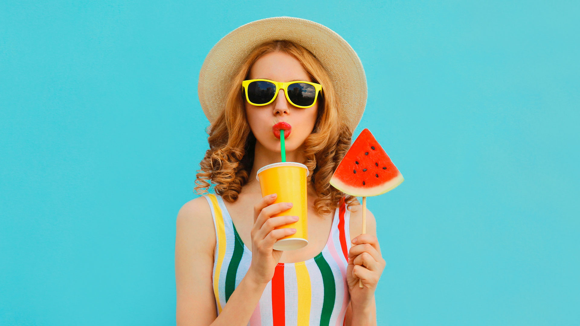 Frau mit Sonnenhut trinkt aus Becher mit Strohhalm, hält Wassermelone.