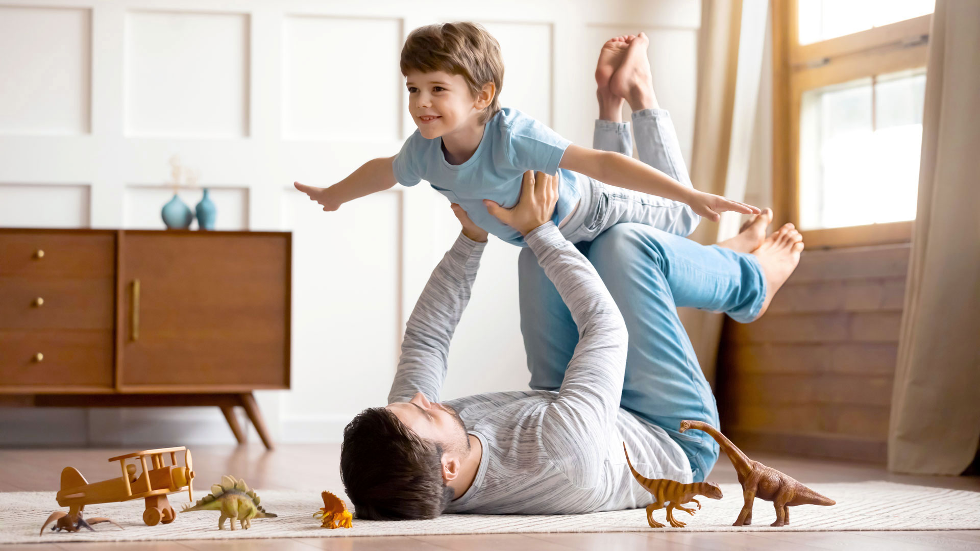 Kind liegt auf dem Rücken eines Erwachsenen, beide spielen mit Spielzeugdinosauriern.