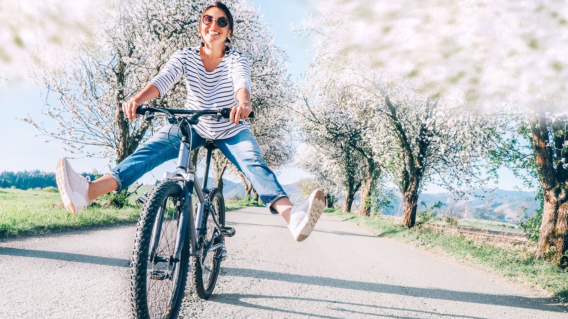 Frau fährt Fahrrad auf Landstraße unter blühenden Bäumen.