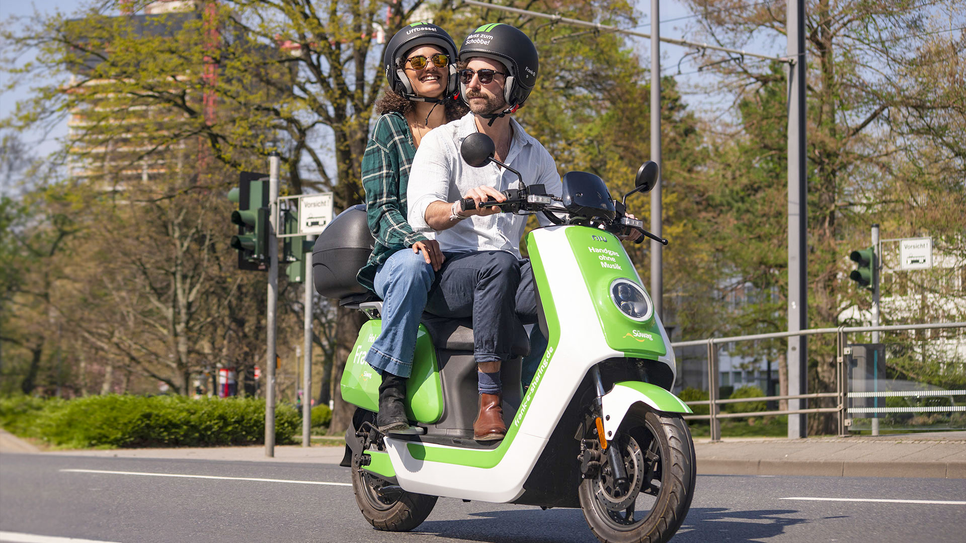 Zwei Personen fahren auf einem grünen Elektroroller durch die Stadt.